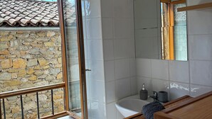 Une salle de bain avec une baignoire, un lavabo, un miroir et une fenêtre donnant sur un mur en pierre et un toit recouvert de tuiles.