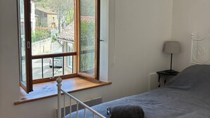 Une chambre avec une grande fenêtre, un lit, une table de chevet avec une lampe, et une vue sur la rue et les bâtiments environnants.