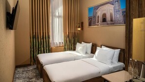 A hotel room with two beds, a large window with curtains, a wall-mounted TV, and a painting of a building.