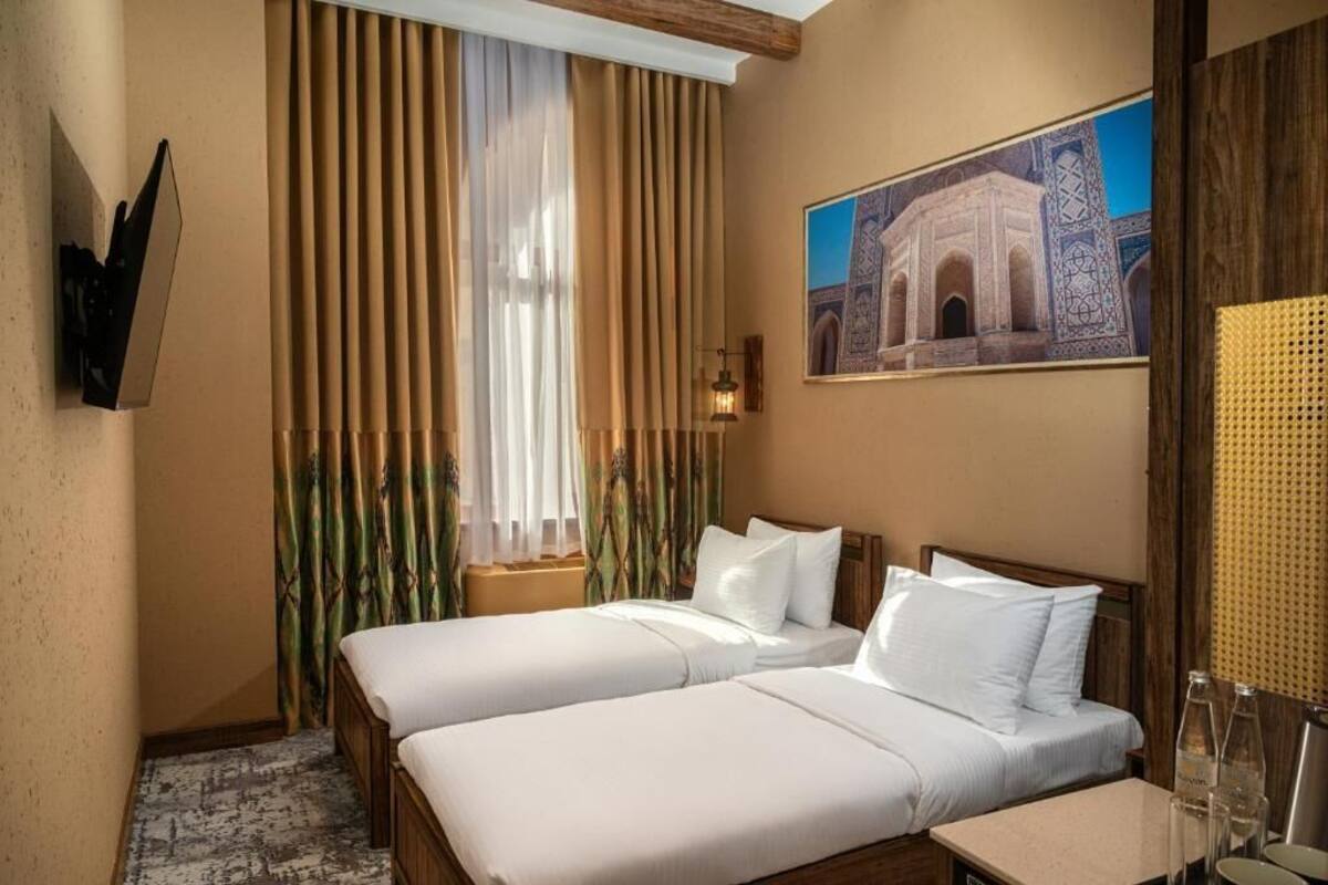 Ein Hotelzimmer mit zwei Betten, einem großen Fenster mit Vorhängen, einem an der Wand befestigten Fernseher und einem Gemälde eines Gebäudes.
