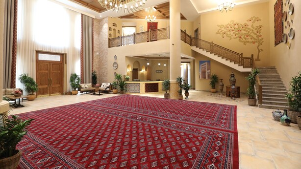 A spacious lobby with a red patterned carpet, a chandelier, a staircase, and multiple potted plants.