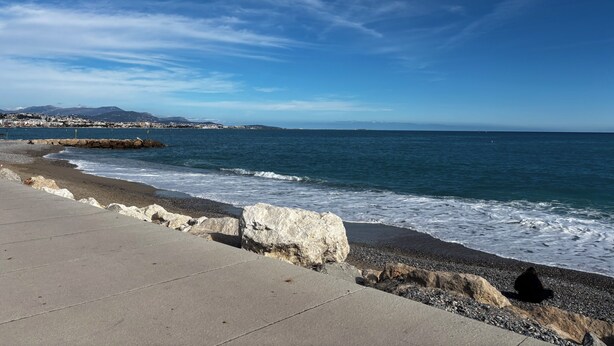 Une promenade en bord de mer, avec une allée en béton, une côte rocheuse et une vue sur la mer et les montagnes lointaines.