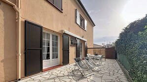 Une maison avec une terrasse en pierre, un coin repas avec des chaises et une haie.