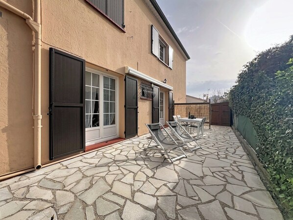 A house with a stone patio, a dining area with chairs, and a hedge.