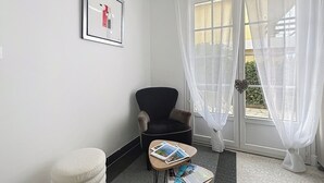 Une pièce comprenant un fauteuil noir, une petite table avec des magazines, un pouf blanc et une fenêtre ornée de rideaux légers.