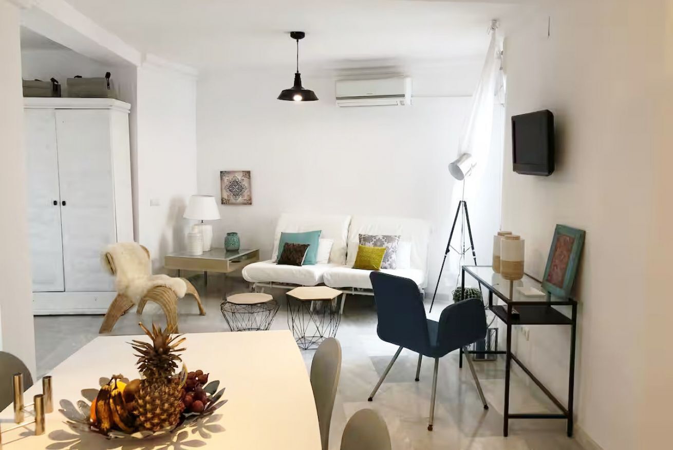 A modern living room with a white sofa, a dining table with fruit, a white cabinet, and a TV mounted on the wall.