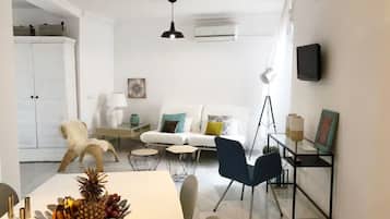 A modern living room with a white sofa, a dining table with fruit, a white cabinet, and a TV mounted on the wall.