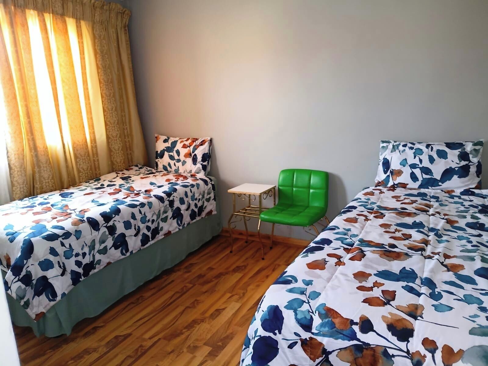 Une chambre avec deux lits simples, un fauteuil vert, une petite table et une fenêtre avec des rideaux.