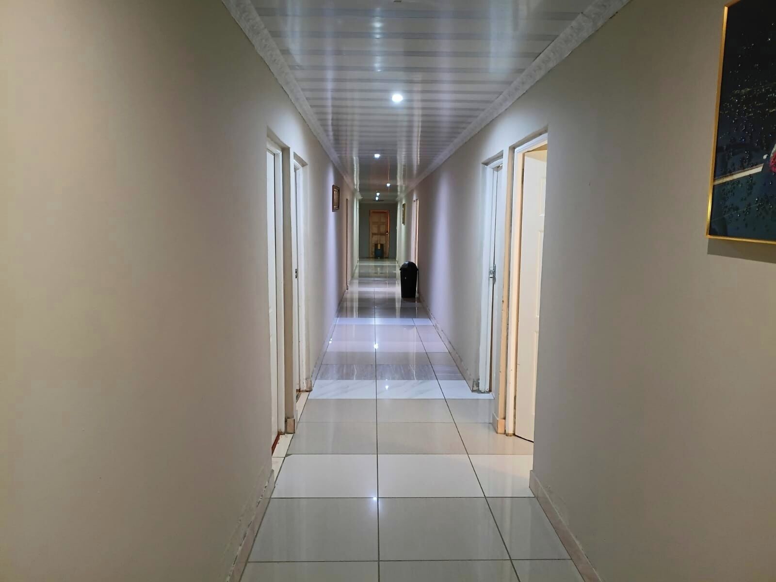 Um corredor comprido e estreito, com chão de azulejos brancos e portas em ambos os lados.