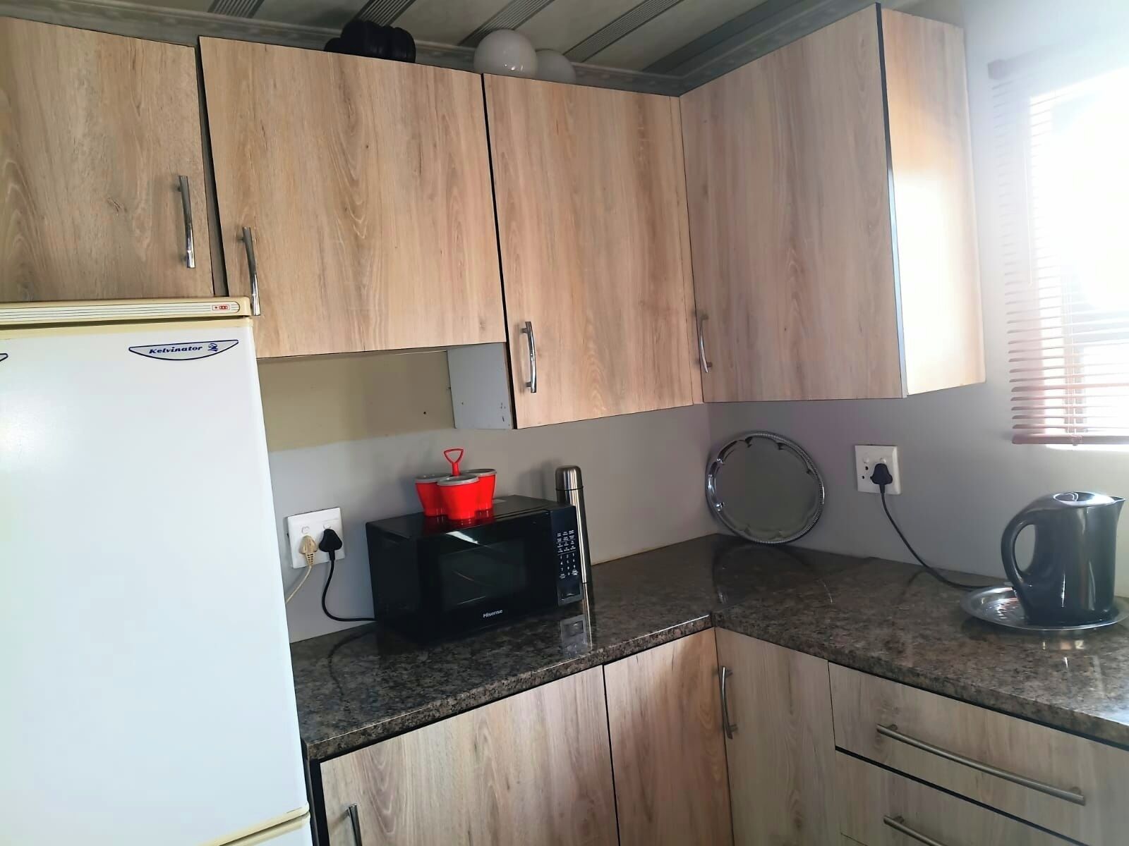 Une cuisine avec des meubles de rangement en bois, un réfrigérateur blanc, un four à micro-ondes, une bouilloire et une assiette ronde posée sur le plan de travail.