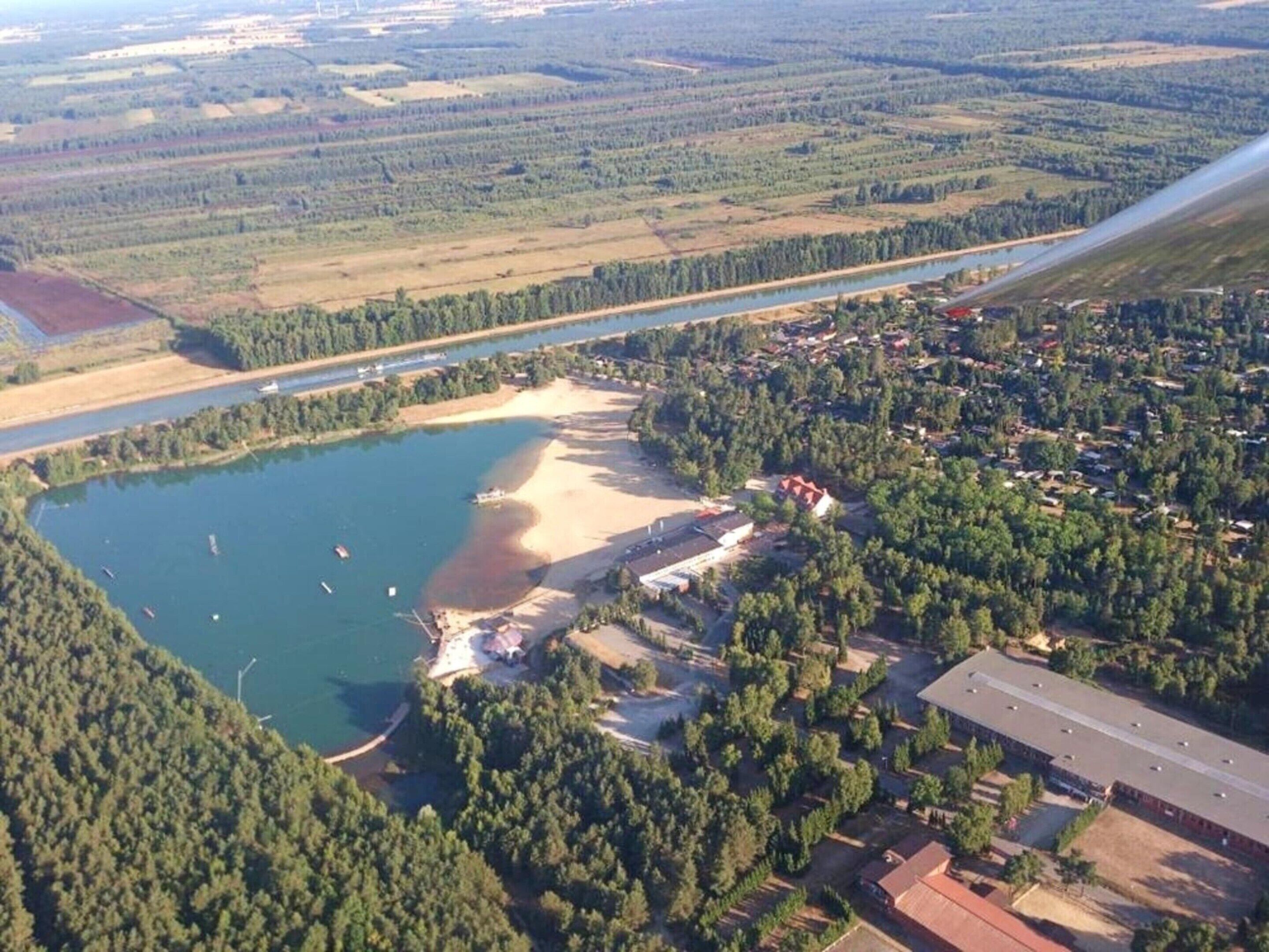 Vue aérienne d’une propriété au bord du lac, avec un petit quai, une plage de sable et une forêt dense qui entoure les lieux.