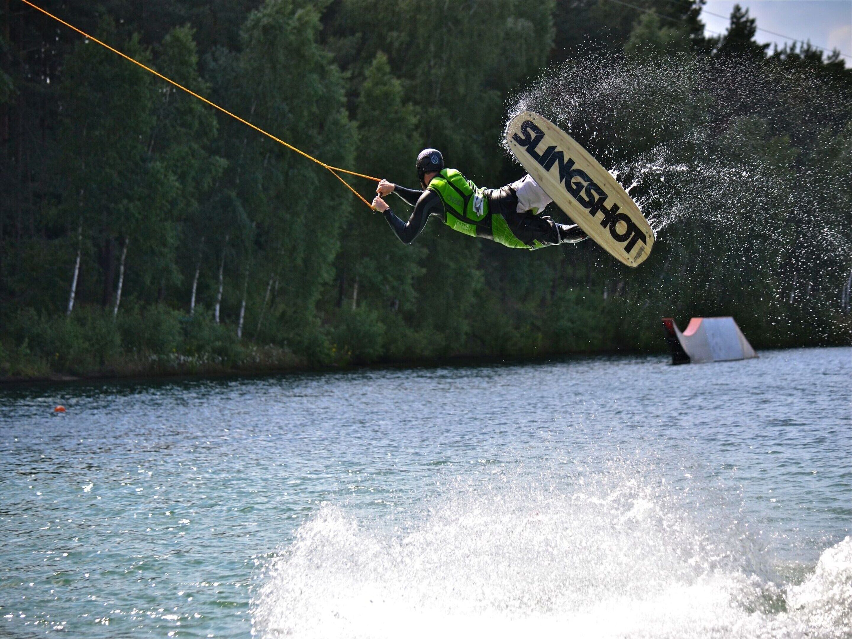 A wakeboarder performing a jump over water with a wakeboard labeled "SLINGSHOT."
