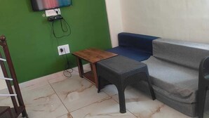 A room with a green accent wall, a flat-screen TV mounted on it, a blue sofa, a small wooden table, a wicker stool, and a marble-tiled floor.