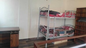 A bunk bed with patterned bedding, a wooden desk, and a bookshelf in a room with plain walls and wooden flooring.