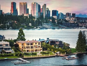 A waterfront property with a large house, a dock, and a city skyline in the background. - Executive Wellness Retreat | Near Lumen Field | Sauna, Gym, Lake Access (Medina)