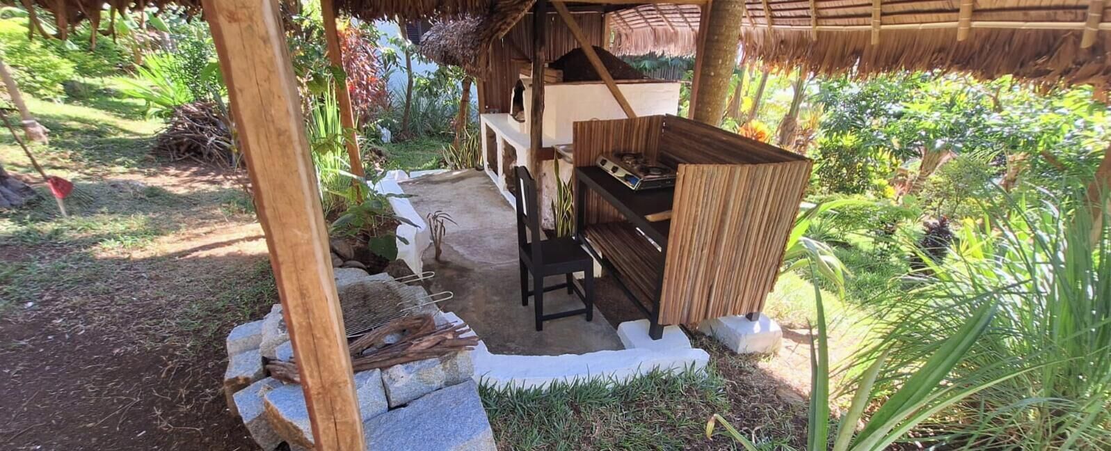 A rustic outdoor seating area with a thatched roof, stone seating, and a small table with a grill.
