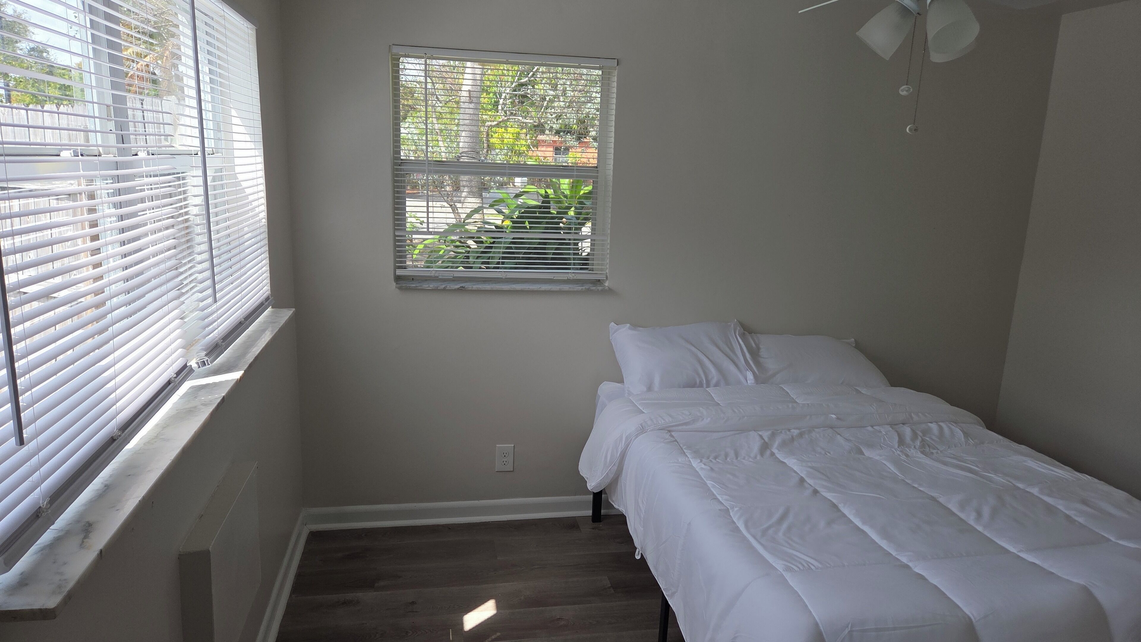 Een kleine kamer met een enkel bed, een raam, een plafondventilator en een stopcontact.