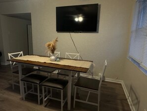 A dining area with a table, chairs, a TV mounted on the wall, and a window with blinds.