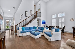 A spacious living room with a large sectional sofa, a wooden staircase, and a dining area. - Emerald Escape: Steps to private beach, Free golf cart, Pool, Luxury Home (Miramar Beach)