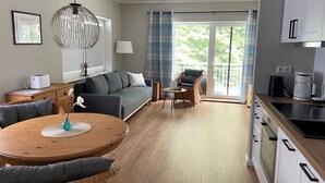 A modern living room with a sofa, a dining table, a wicker chair, a lamp, a coffee maker, and a view of the outdoors through a sliding glass door. - Seeperle by the Lake Vacation Rental (Lalendorf)