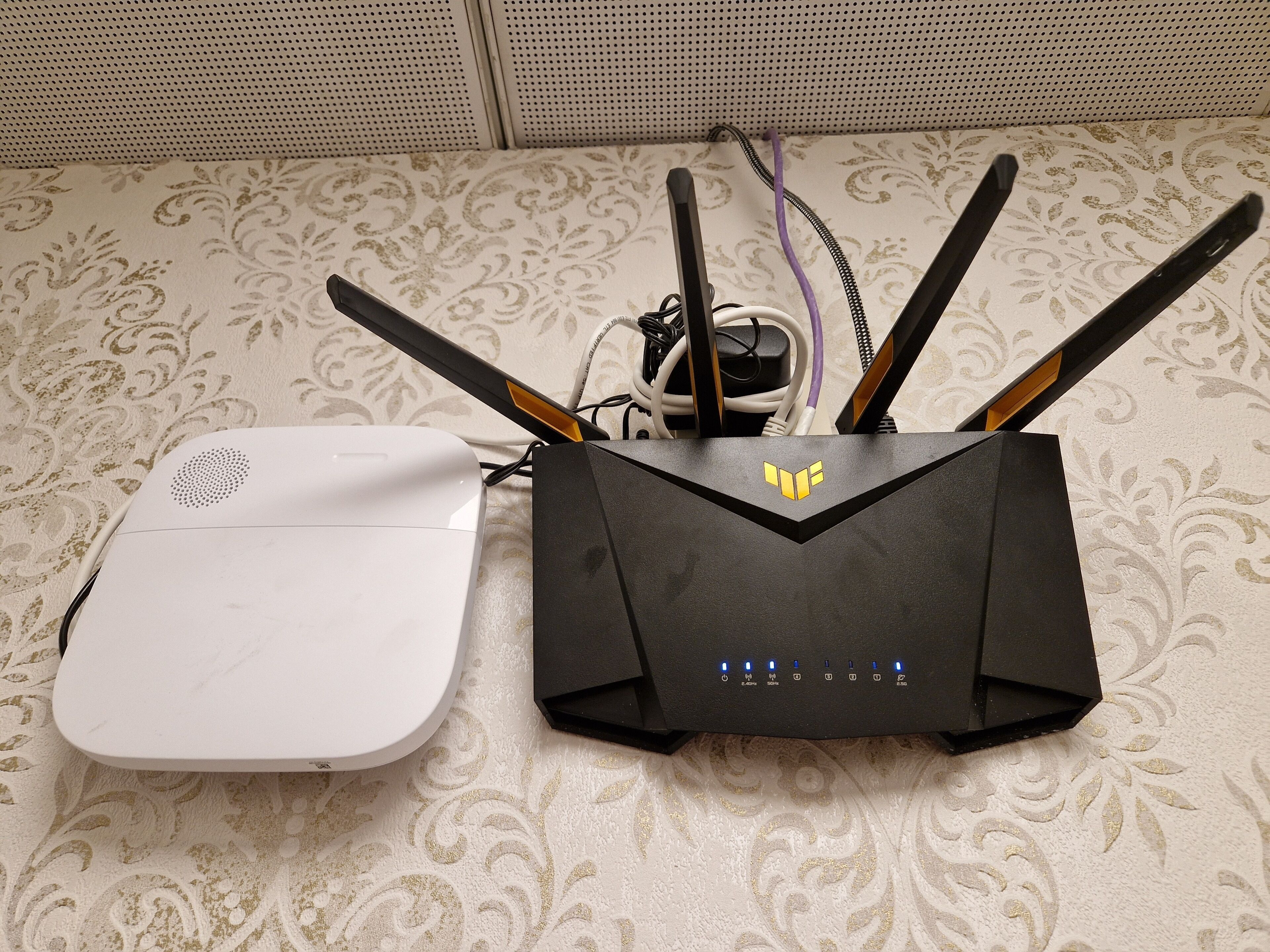 Un dispositivo blanco conectado a un router negro con múltiples antenas sobre una superficie textil estampada.
