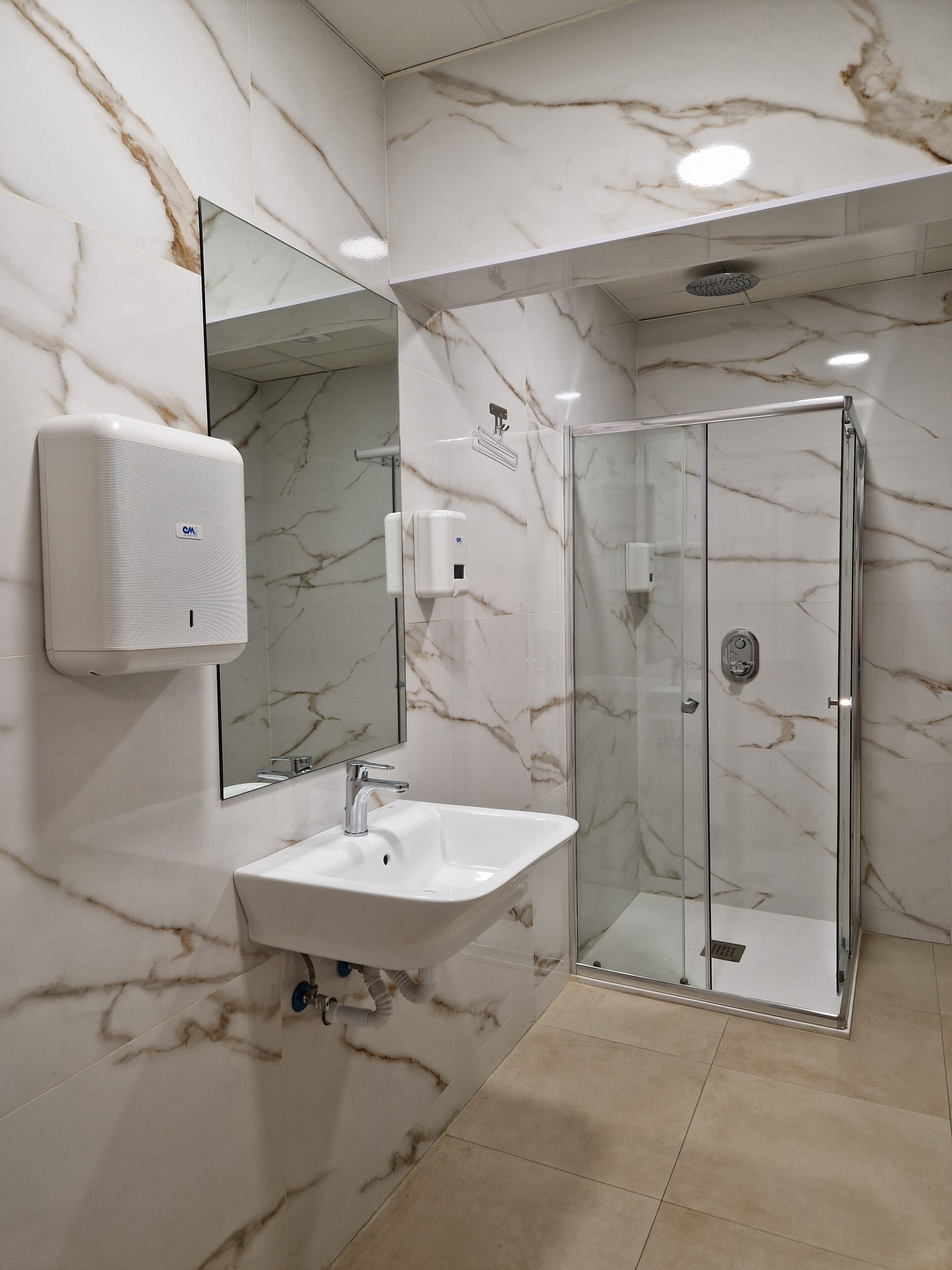 Un baño con pared de mármol, un lavamanos, un espejo, un secador de manos y una ducha con cerramiento de vidrio.