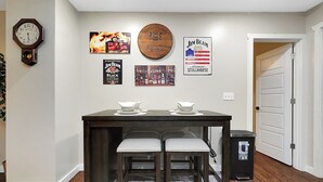 Ein Essbereich mit Tisch, zwei Stühlen, einer Uhr sowie Wanddekorationen, darunter eine Uhr, ein Wandbild mit Feuerstelle, ein Stillhouse-Schild und ein Poster mit Flaschen.