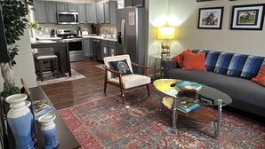 Ein Wohnzimmer mit einer Couch, einem Couchtisch und einem Teppich. Im Hintergrund ist eine Küche mit Schränken und Geräten zu sehen.
