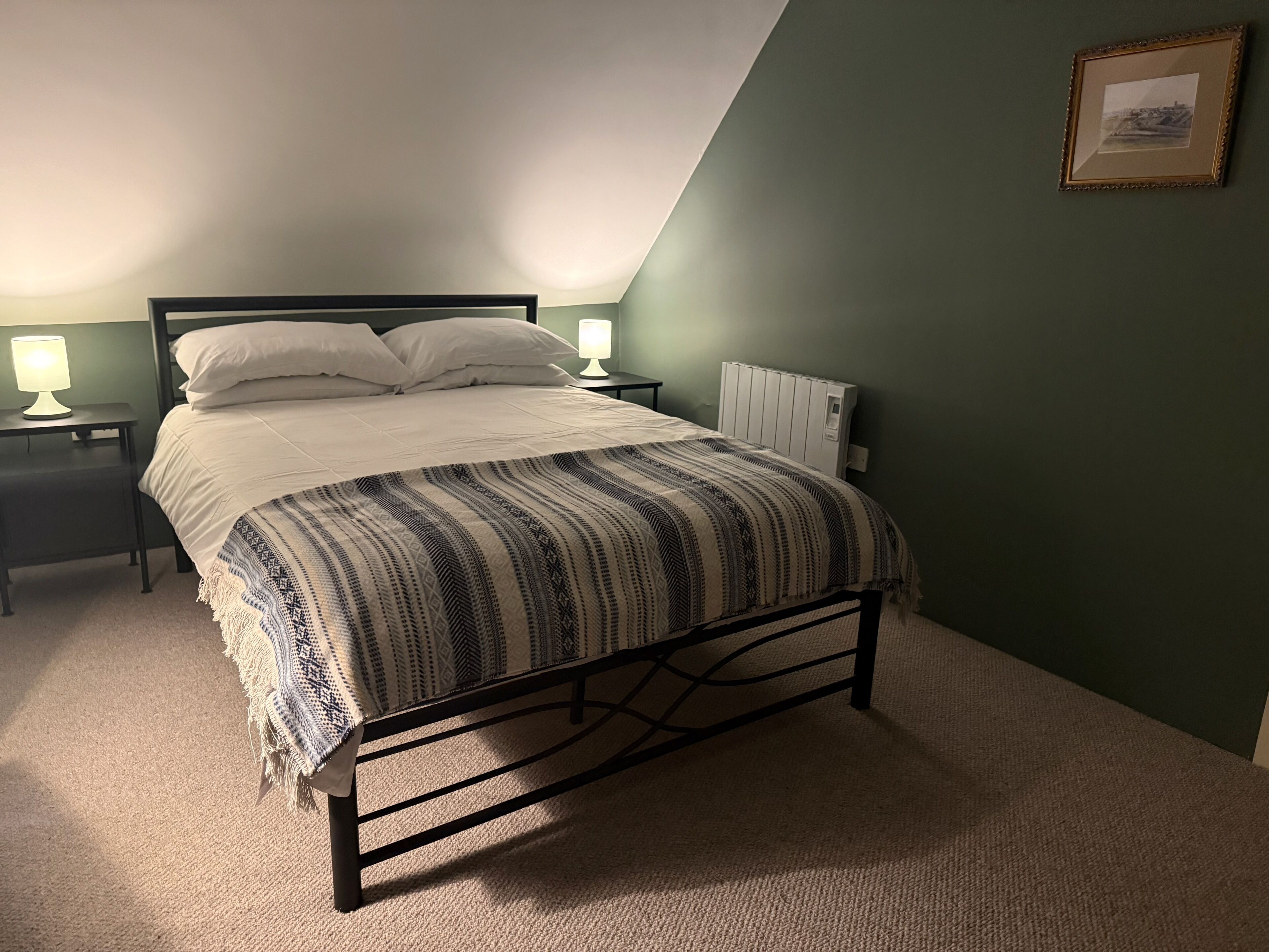 A bedroom with a bed, two bedside tables with lamps, a radiator, and a framed picture on the wall.