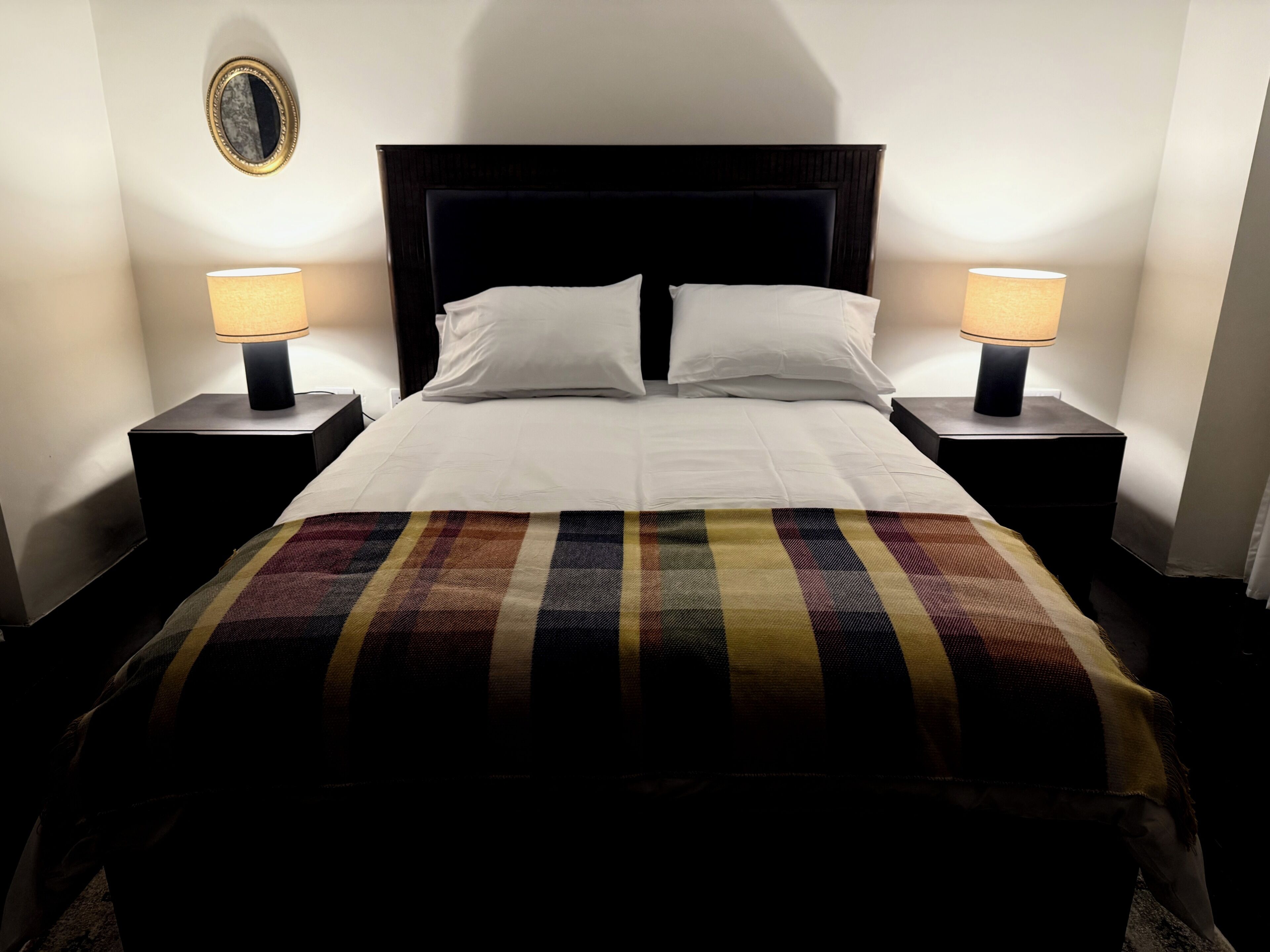 Una cama con sábanas blancas, cabecera oscura y manta a rayas. Hay dos lámparas de noche a ambos lados de la cama.