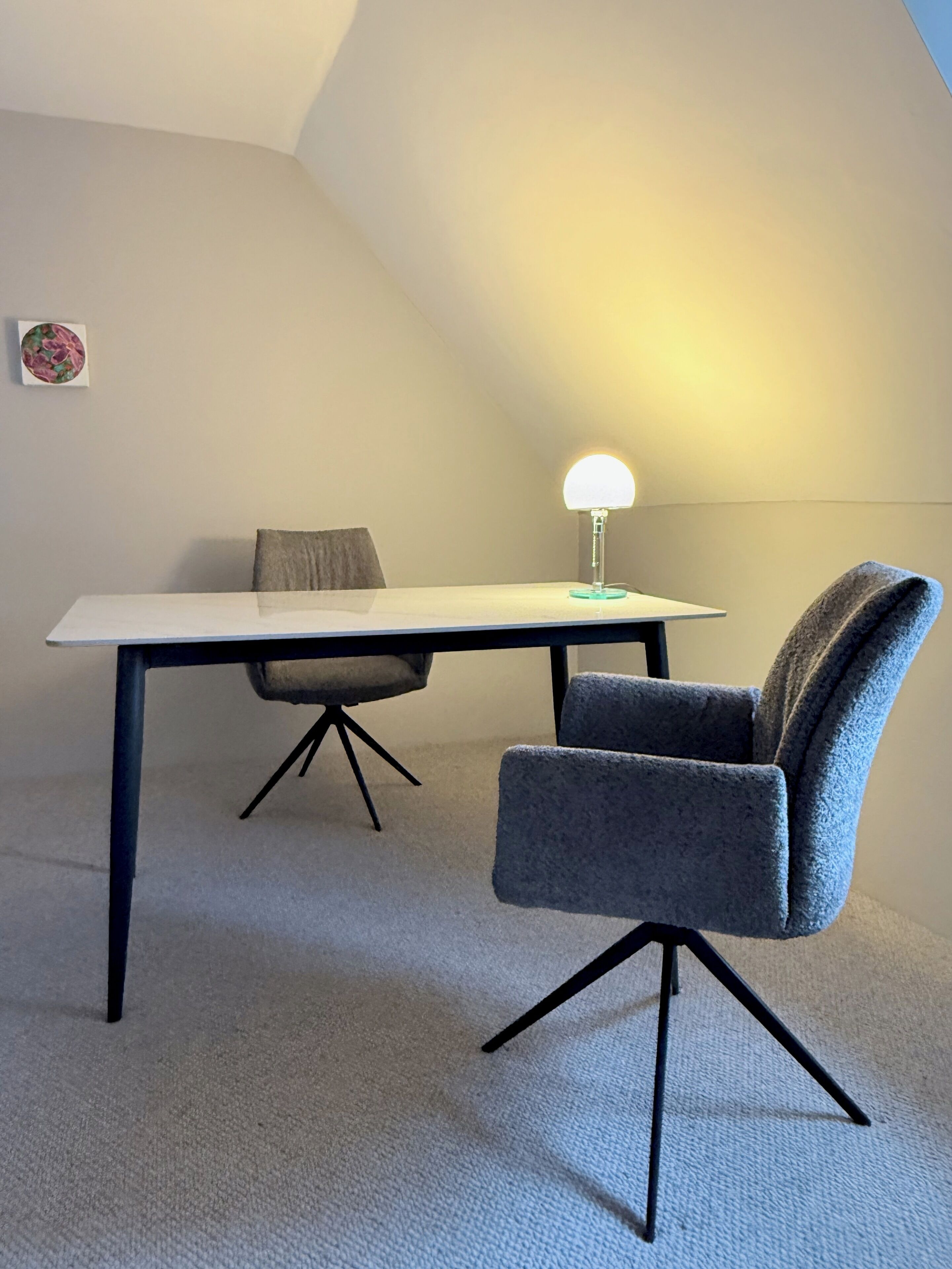 Une pièce avec une table blanche, une chaise grise, une lampe et un sol recouvert d’une moquette.