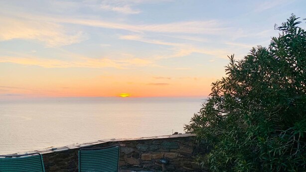 Eine Steinterrasse mit zwei grünen Stühlen, einer Steinsäule und Blick auf das Meer bei Sonnenuntergang.