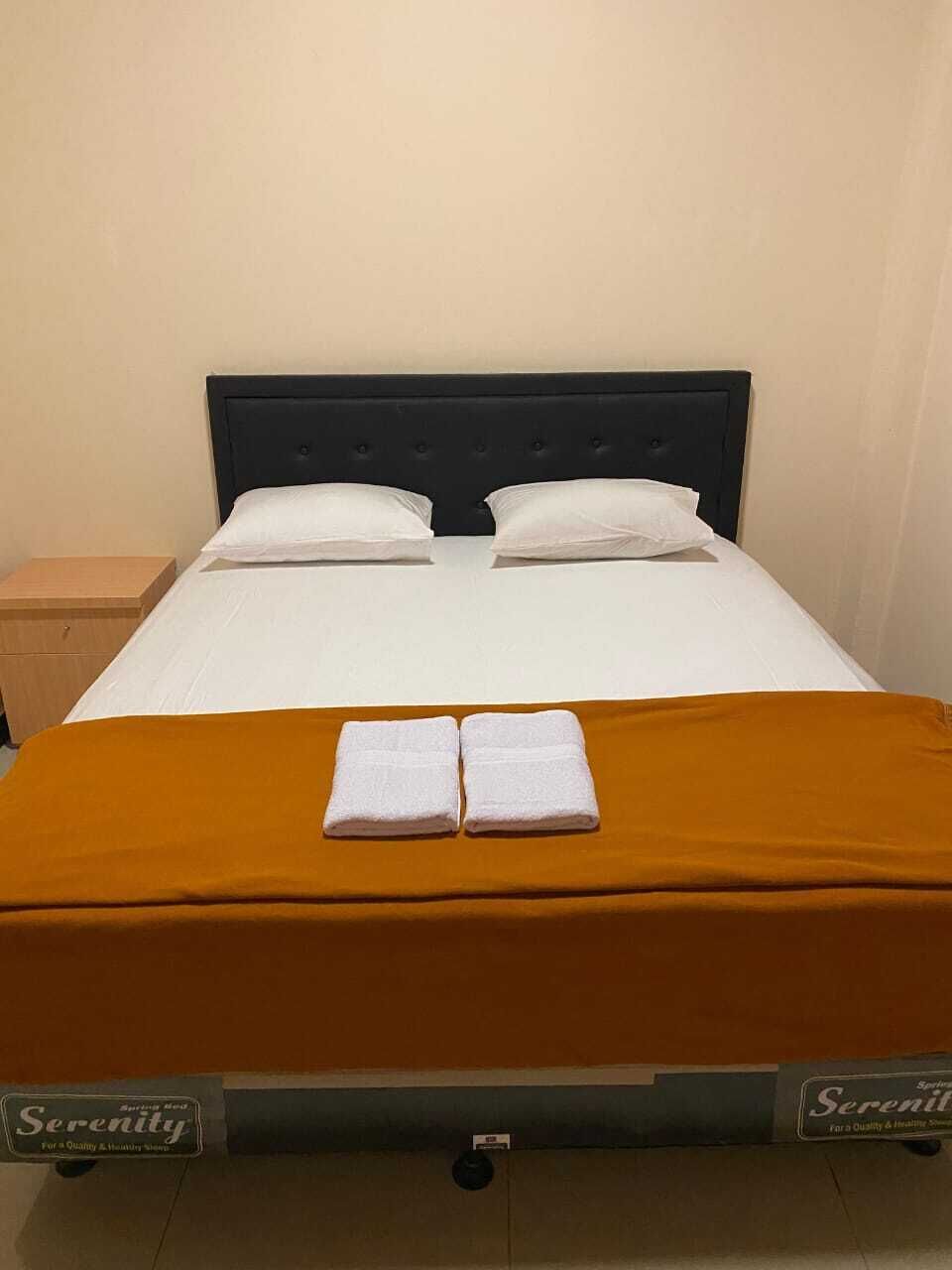 Una cama individual con cabecera oscura, ropa de cama blanca y una colcha naranja. Hay dos toallas dobladas sobre la cama.