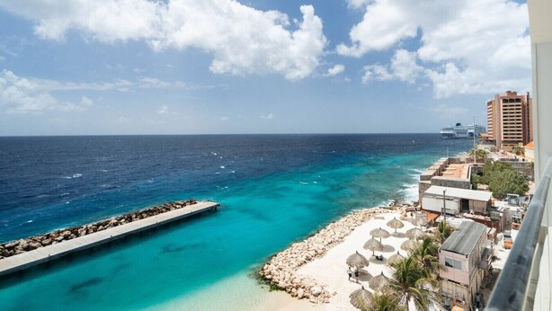 Vista costera con un muelle, aguas turquesas cristalinas, una playa arenosa y un paisaje urbano con edificios y un crucero en la distancia.