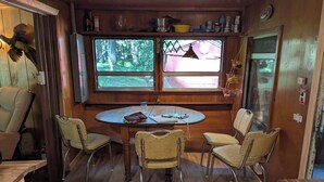 A cabin interior with a round wooden table, four chairs, a window with a view of a forest, and a shelf with various items.