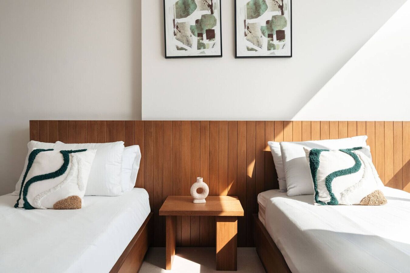 Ein Hotelzimmer mit zwei Betten, Holz-Kopfteilen, weißer Bettwäsche, einem kleinen Nachttisch mit einer Vase und abstrakter Kunst an der Wand.