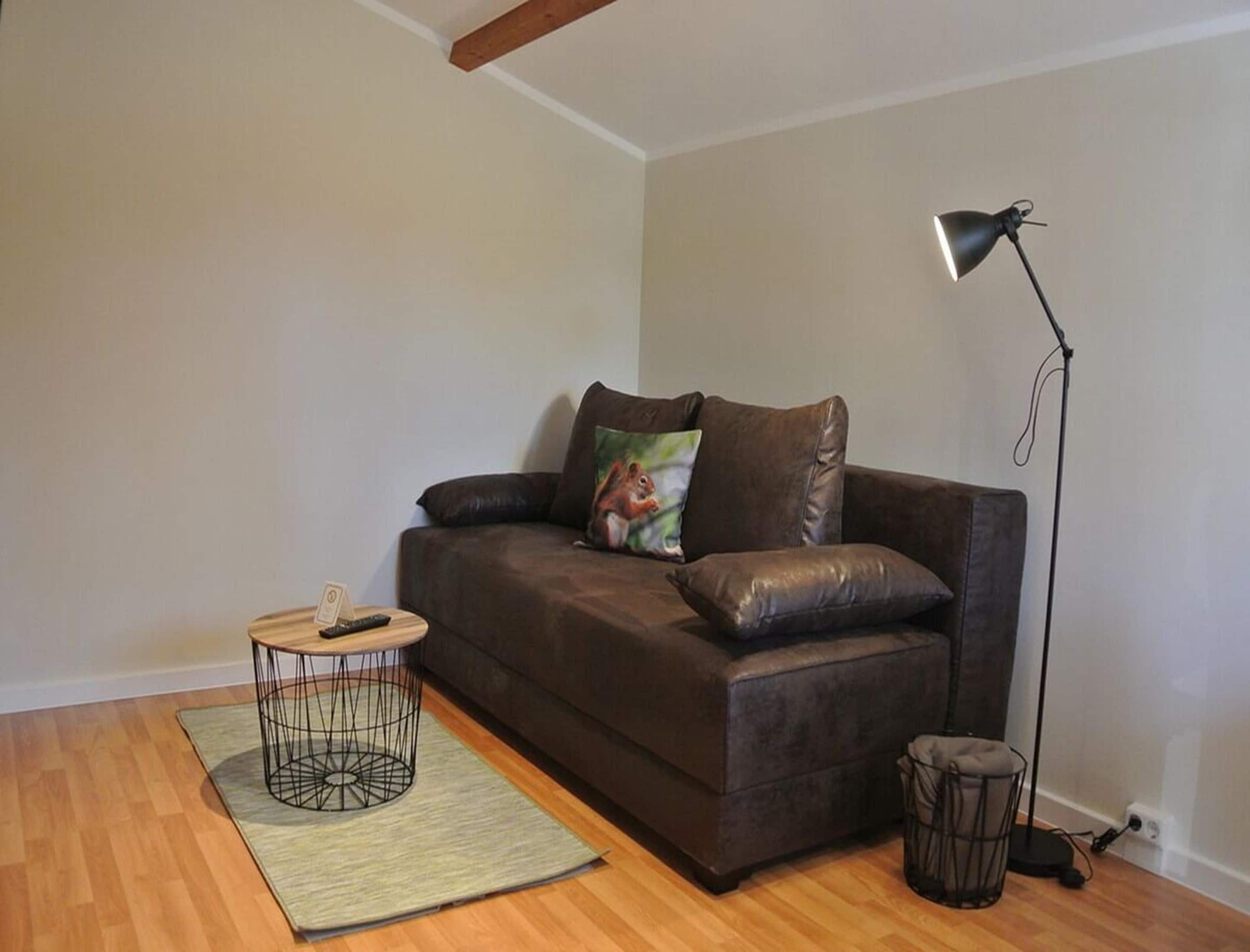 A living room with a brown leather sofa, a small round coffee table, a floor lamp, and a trash bin.