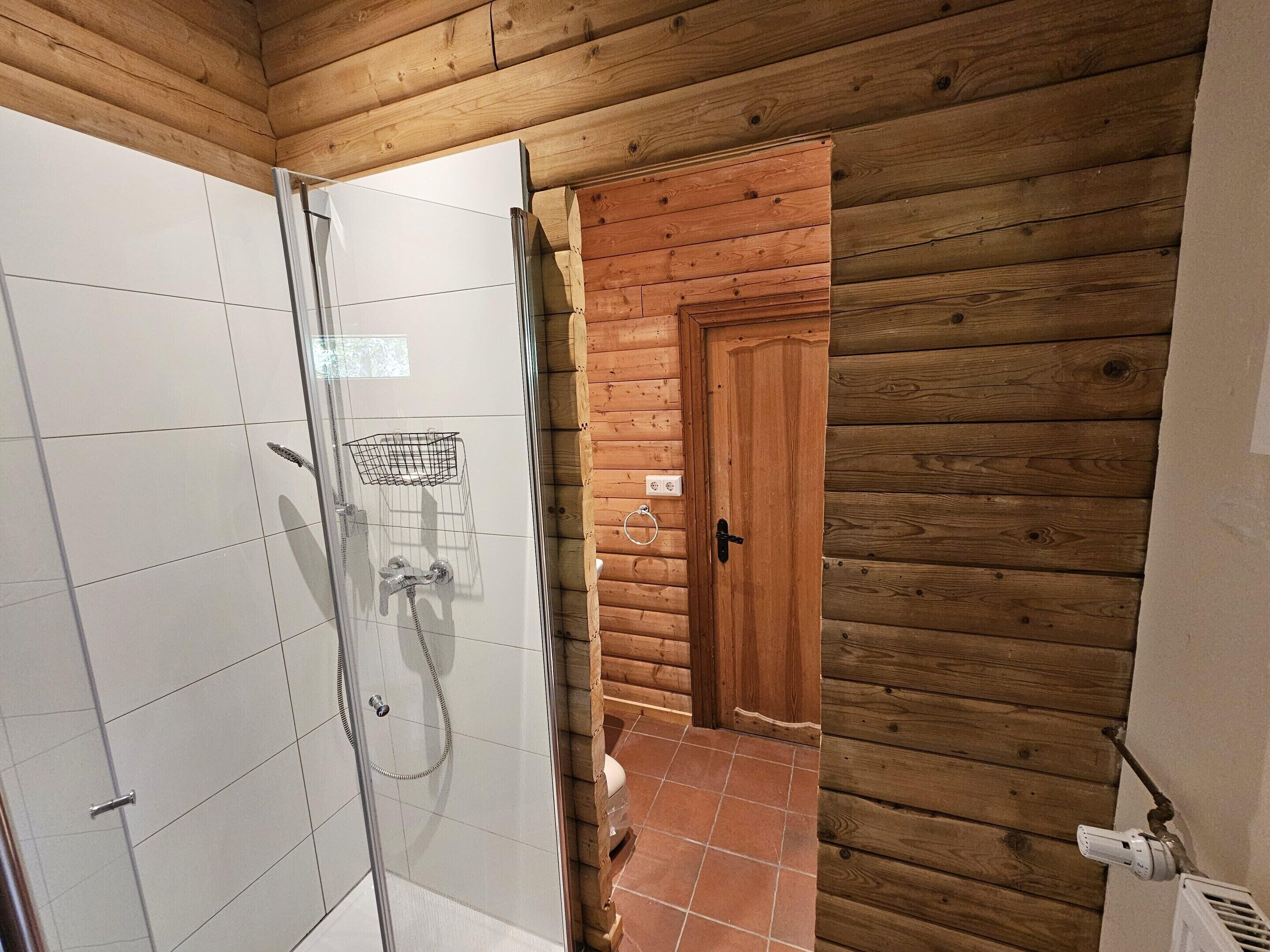 Ein Badezimmer in einer Holzhütte mit einer Duschkabine aus Glas, einer Holztür und einem gefliesten Boden.