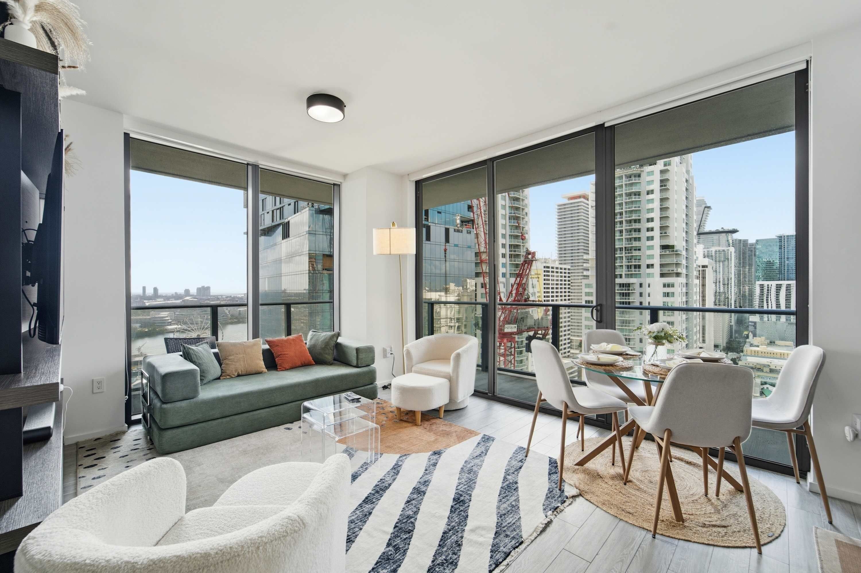 Una sala moderna con un sofá, sillas, una mesa de centro y una mesa de comedor preparada para cuatro comensales. Amplias ventanas ofrecen vistas del paisaje urbano.