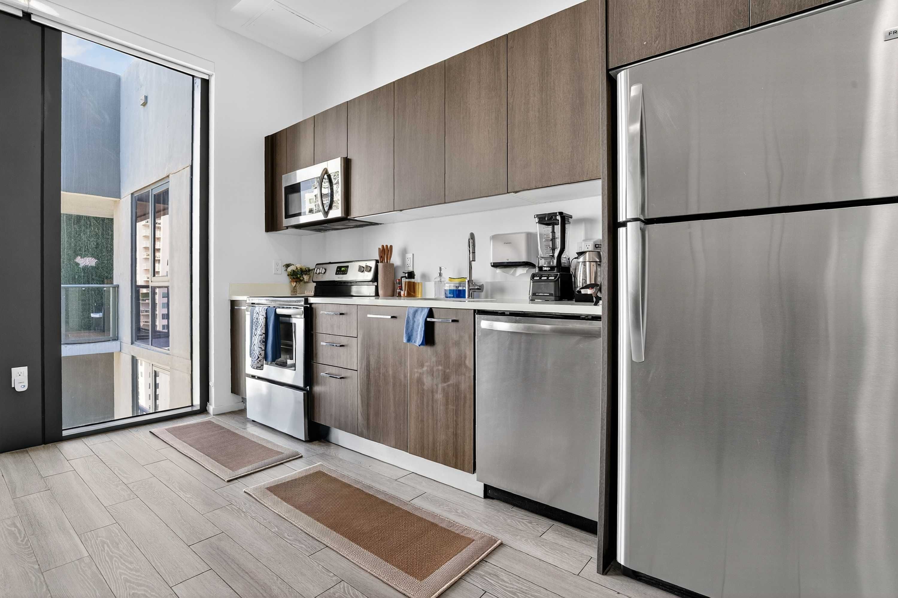 A modern kitchen with stainless steel appliances, wooden cabinets, and a sliding glass door leading to a balcony.