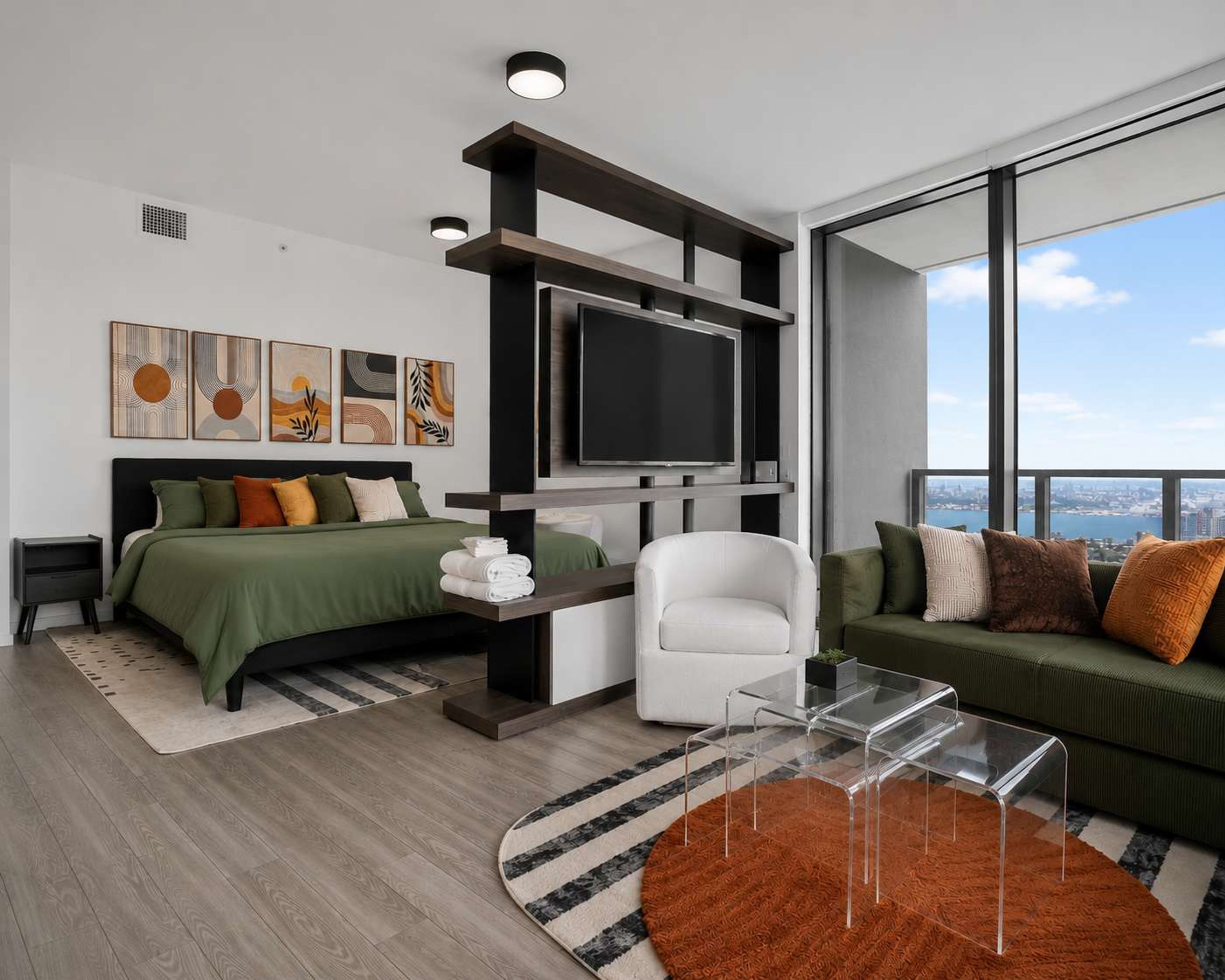 A modern bedroom with a bed, a TV, a sofa, and a chair. There are artworks on the wall and a view of the city through large windows.