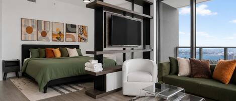 A modern bedroom with a bed, a TV, a sofa, and a chair. There are artworks on the wall and a view of the city through large windows.