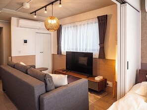 Moderni hotellihuone, jossa on sohva, televisio, pieni pöytä ja kattovalaisin.