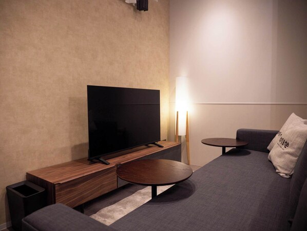 Ein modernes Wohnzimmer mit Flachbildfernseher, einem Holztisch, einem runden Couchtisch, einem grauen Sofa und einem kleinen Beistelltisch mit Lampe.