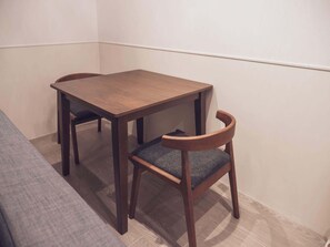 Ein kleiner, ordentlich eingerichteter Raum mit einem Holztisch, zwei Stühlen, einem grauen Sofa und einer schlichten Wand.