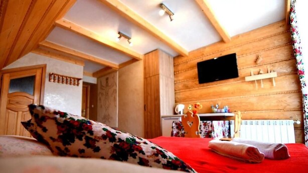 A hotel room with a wooden ceiling, a bed with floral bedding, a red blanket, and a mounted television. - Pokoje Gościnne Majerczyk  (Zakopane)