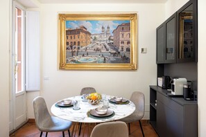 A dining area with a round table, chairs, a painting, and a kitchen area with appliances. - Modern 2-bedroom apartment Spanish Steps • Trinity Domus (Roma)