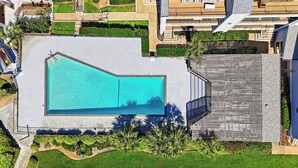 Vista aérea de una piscina rodeada de jardines, una terraza de madera y un edificio al fondo.