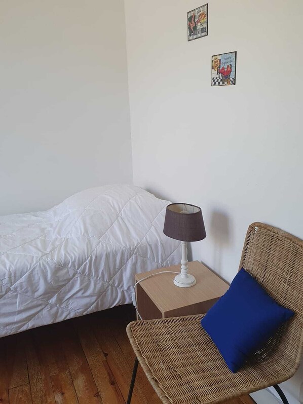 Un dormitorio con una cama, una mesita con lámpara, una silla de mimbre con almohada azul y dos adornos en la pared.
