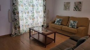 Uma sala de estar com um sofá marrom, uma mesa de centro de vidro e uma cortina estampada.